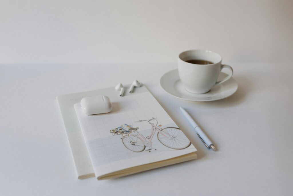 A minimalist setup with a cup of coffee, notepad, pen, and wireless earbuds on a white background.